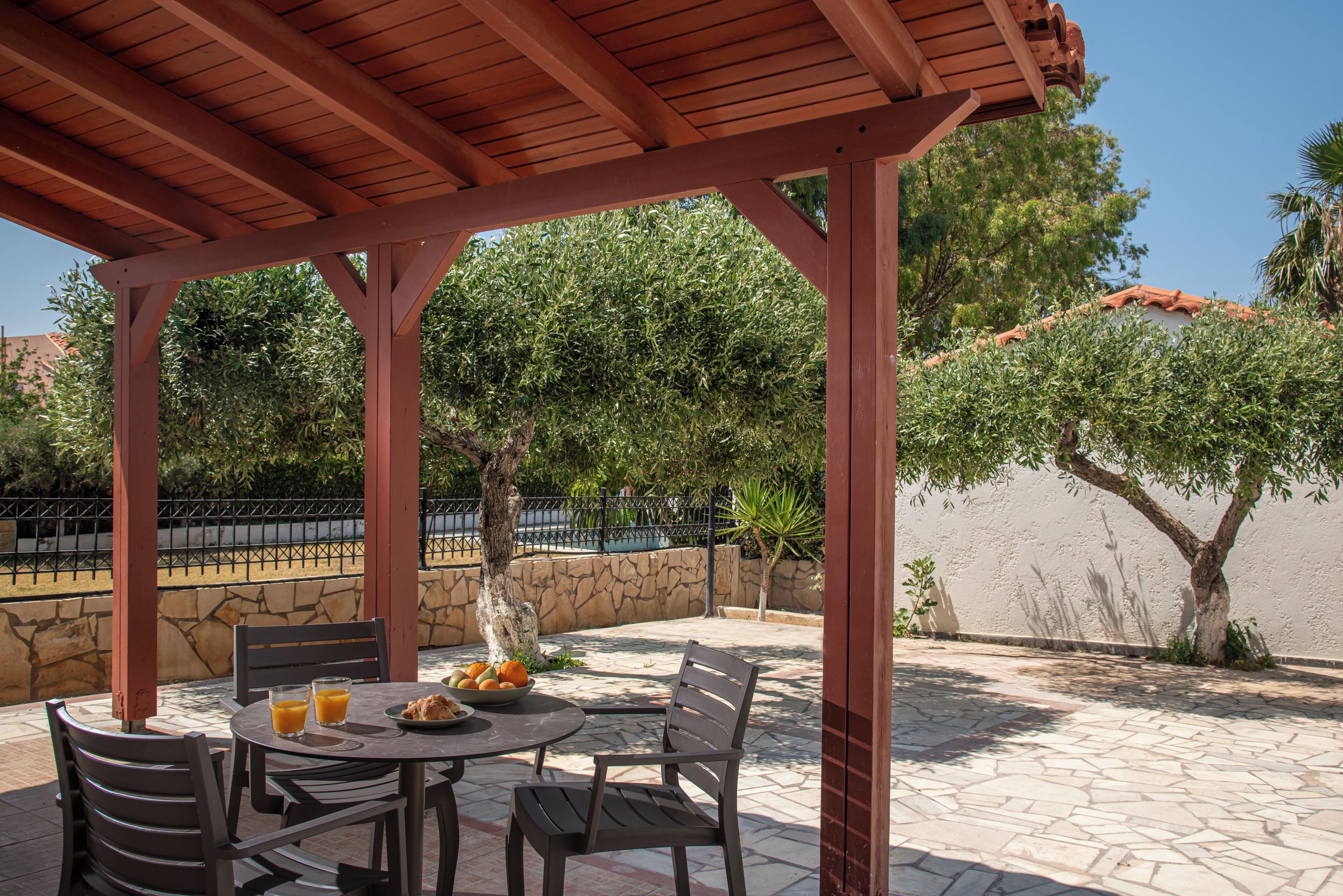Un patio al aire libre con una pérgola de madera, suelo de piedra, una mesa con sillas y un cuenco de fruta.