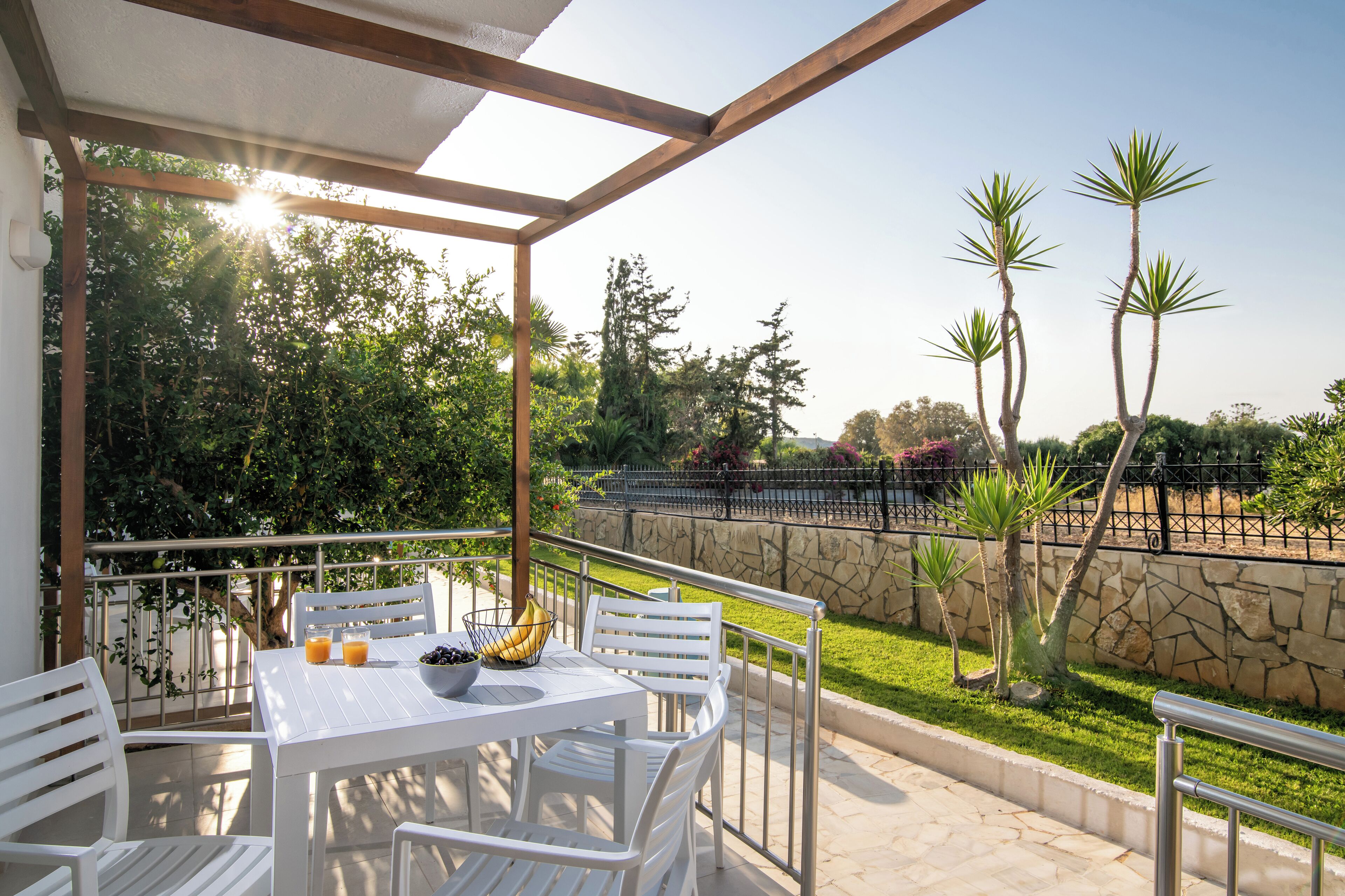 Un área de estar al aire libre techada con una mesa y sillas, un muro de piedra y vistas a un jardín con palmeras.