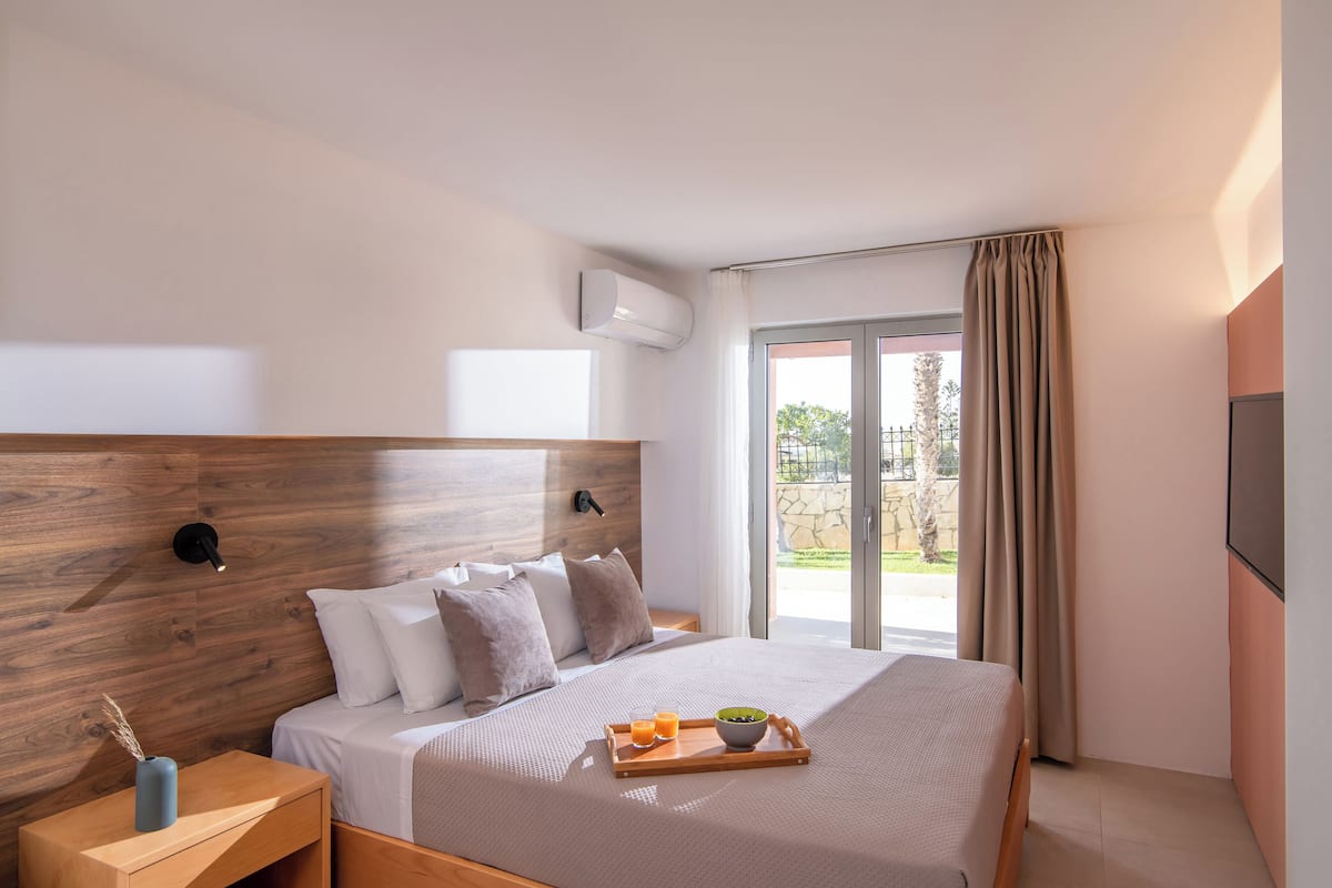 Una habitación de hotel moderna con cabecera de madera, cama con ropa blanca, una bandeja con productos de desayuno y una puerta corrediza de vidrio al exterior.