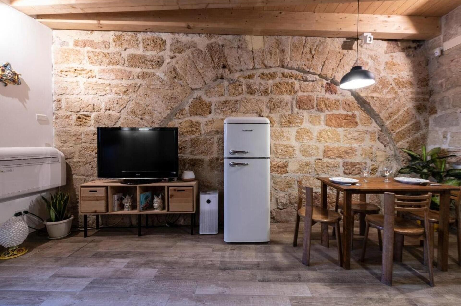 A compact kitchen with a stone wall, a flat-screen TV, a white refrigerator, wooden furniture, and a dining table.