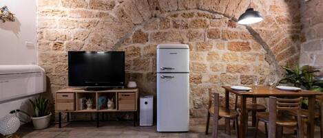 A compact kitchen with a stone wall, a flat-screen TV, a white refrigerator, wooden furniture, and a dining table.
