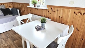 Une pièce chaleureuse avec une table blanche, des chaises, une plante en pot et une petite étagère ornée d’objets décoratifs.