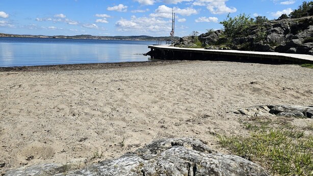 Une plage de sable avec une jetée en béton qui s’avance dans une eau calme, entourée d’un paysage rocheux et d’une végétation clairsemée.