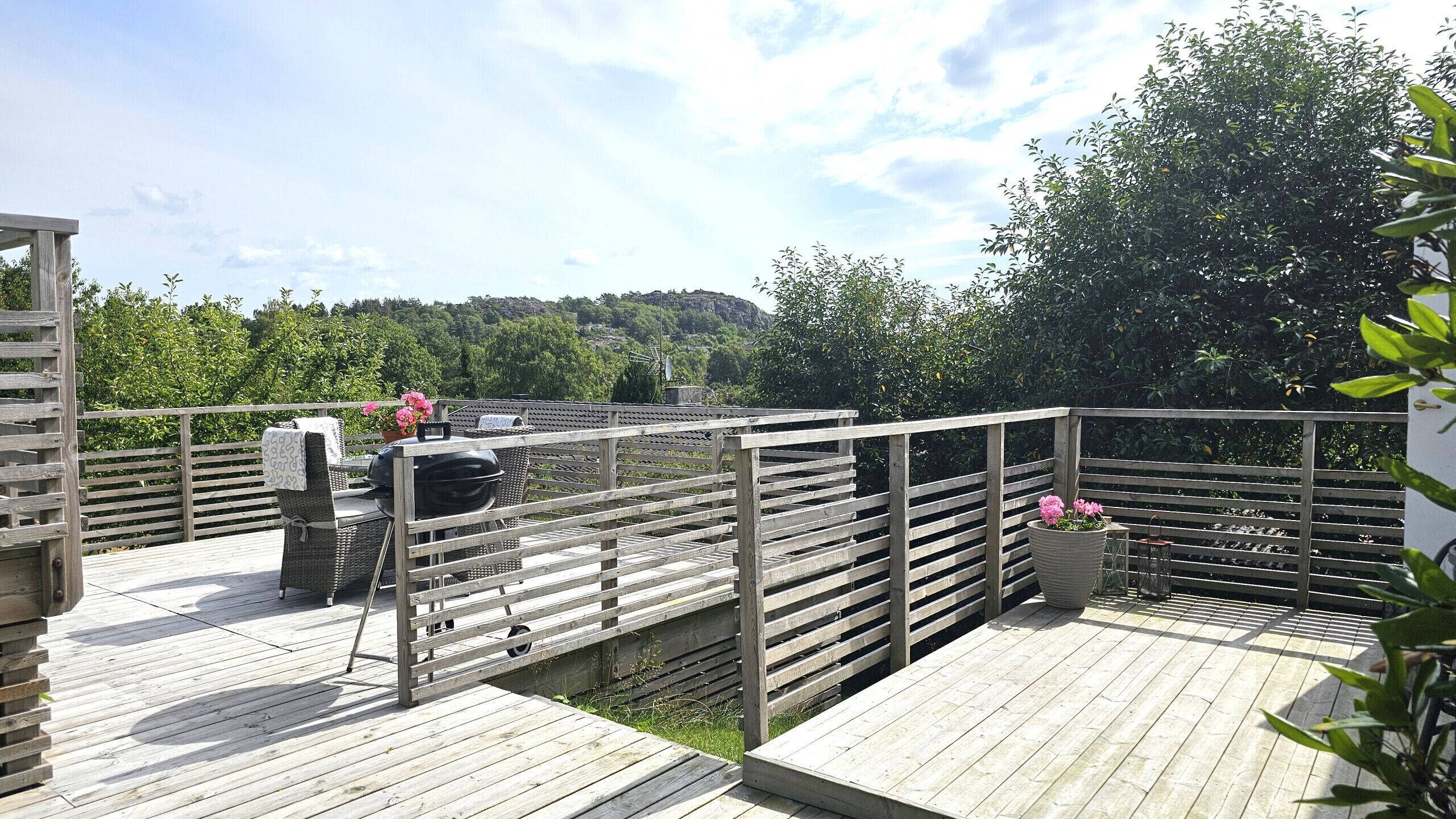 A wooden deck with a grill and seating area, surrounded by greenery and a view of a hilly landscape.