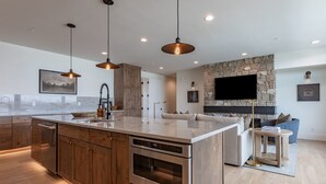 A modern kitchen with a large island, built-in oven, and wooden cabinets. There is a stone fireplace and a flat-screen TV mounted on the wall. - Basin Three Peaks | 8BR Luxury Group Escape (Driggs)