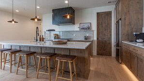 A modern kitchen with a large island, wooden stools, pendant lighting, and a stainless steel refrigerator. - Basin Three Peaks | 8BR Luxury Group Escape (Driggs)