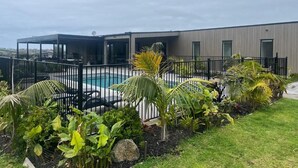 A modern building with a swimming pool surrounded by a black fence and tropical plants. - A Private Resort in Mangawhai Heads (Mangawhai)