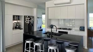 A modern kitchen with a central island, bar stools, a built-in oven, and a large window. - A Private Resort in Mangawhai Heads (Mangawhai)