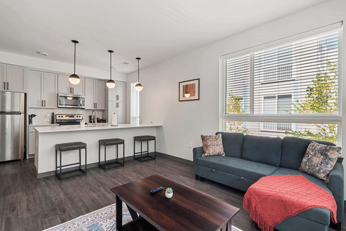 A modern kitchen with white cabinets, a stainless steel refrigerator, and a built-in oven. There are three bar stools and pendant lights above the kitchen island. A gray sofa with a red throw blanket is positioned near a large window with blinds, and a framed picture hangs on the wall.