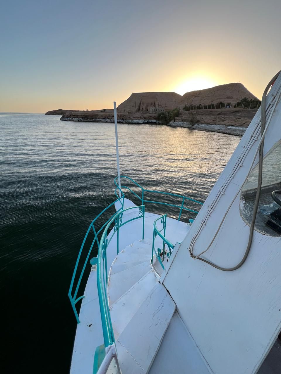 A boat's deck with a metal railing and a view of the sea and a rocky coastline during sunset.