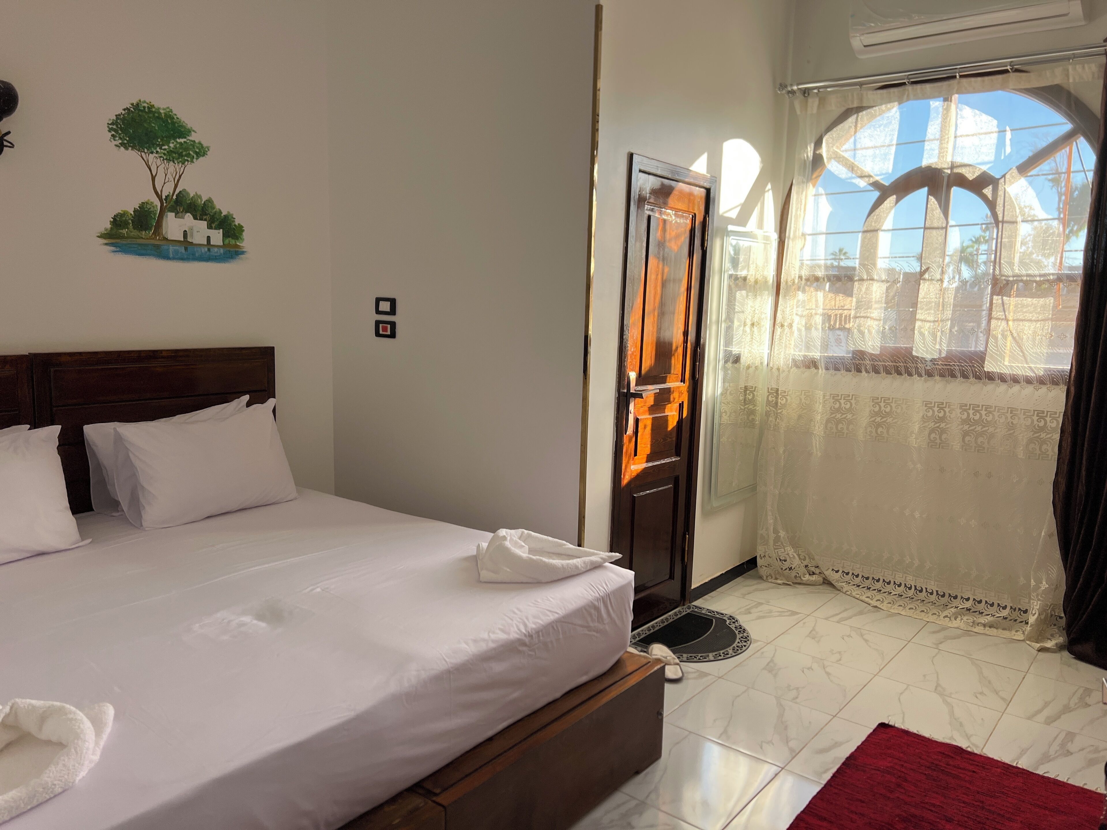 Habitación de hotel con cama, cabecera de madera, ventana que ofrece vista a palmeras y puerta con espejo.