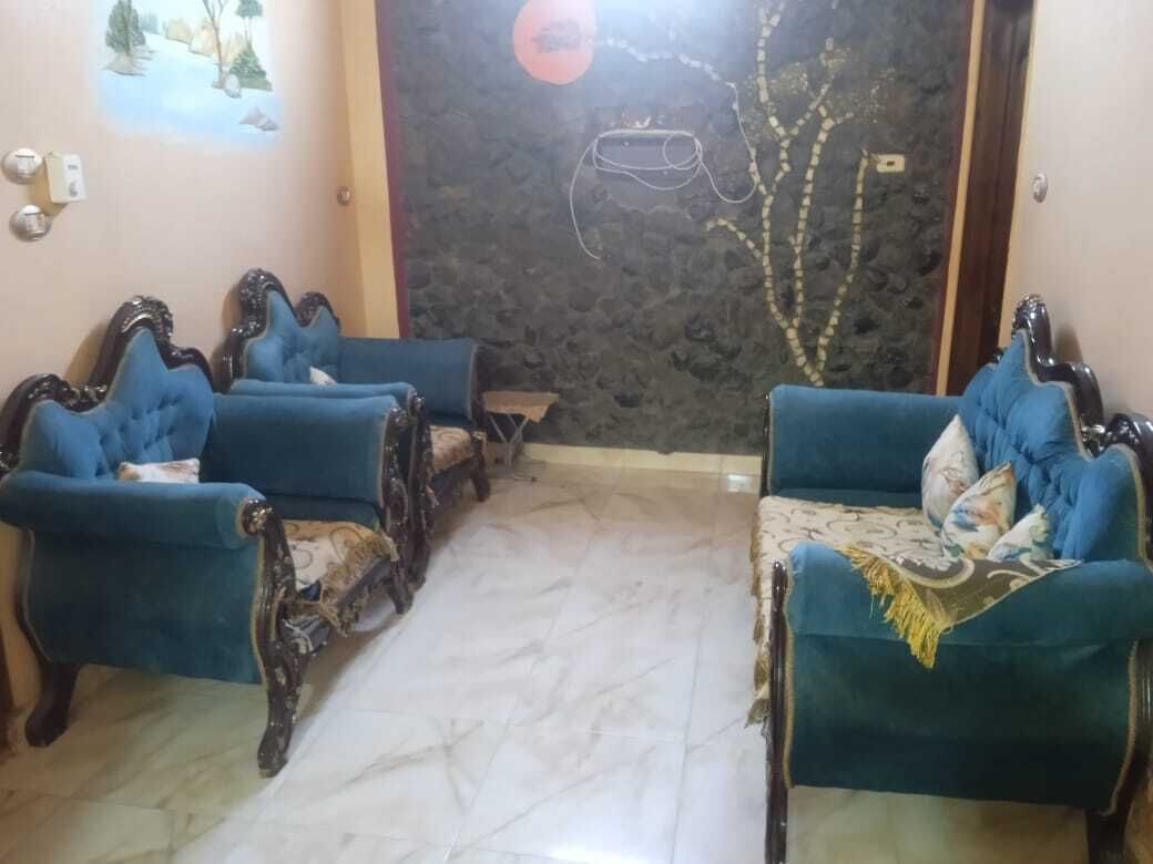 A room with two blue armchairs and a patterned sofa, a marble floor, and a wall with a mural and a hanging light fixture.