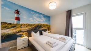 Ett hotellrum med en stor säng, två sängbord, ett litet bord, en lampa, ett fönster med gardiner och en väggmålning som föreställer ett fyrtorn.