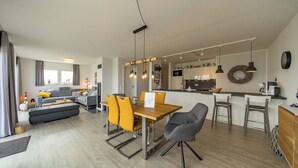 Ett modernt vardagsrum med en soffa, ett matbord och stolar, ett kök i bakgrunden och utsikt över utomhusmiljön.