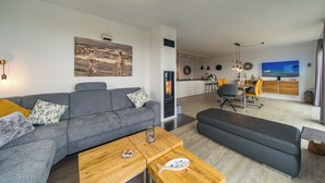 Ett modernt vardagsrum med en grå soffa, ett träbord och en svart pall. På väggen hänger en tavla, det finns en öppen spis och en matplats med gula stolar.