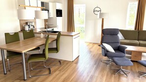 A modern, well-lit living area with a wooden dining table, green chairs, a grey sofa, and a kitchen in the background. - North Sea apartment with sauna & south-facing balcony - sea breeze and beach just 200 m away (Nordseeheilbad Schillig)