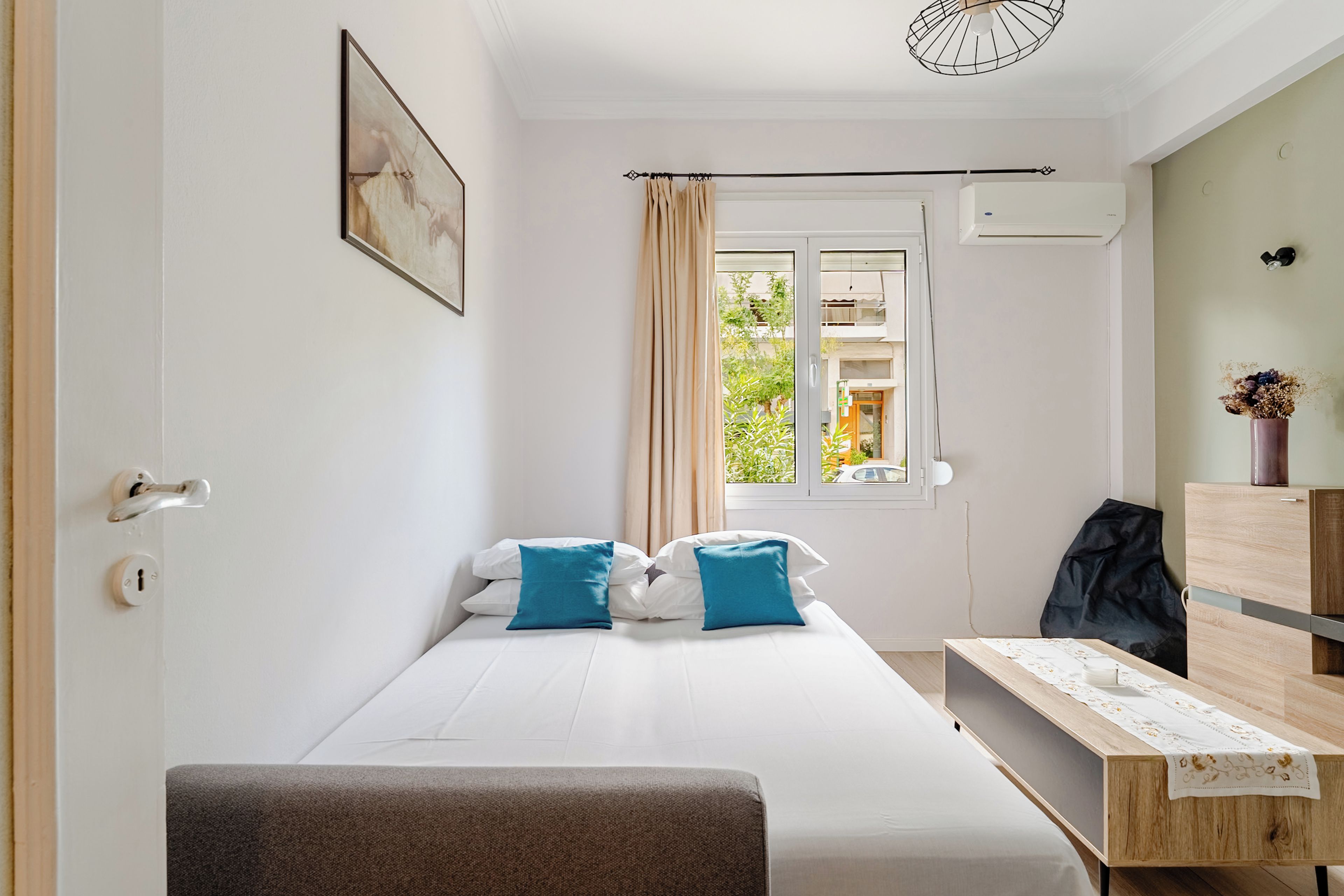 A small, well-lit bedroom with a single bed, a bedside table, a chair, and a window with curtains.