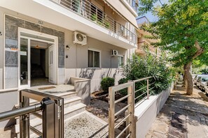 Un edificio de varios pisos con entrada protegida, escalones exteriores y un árbol en primer plano.