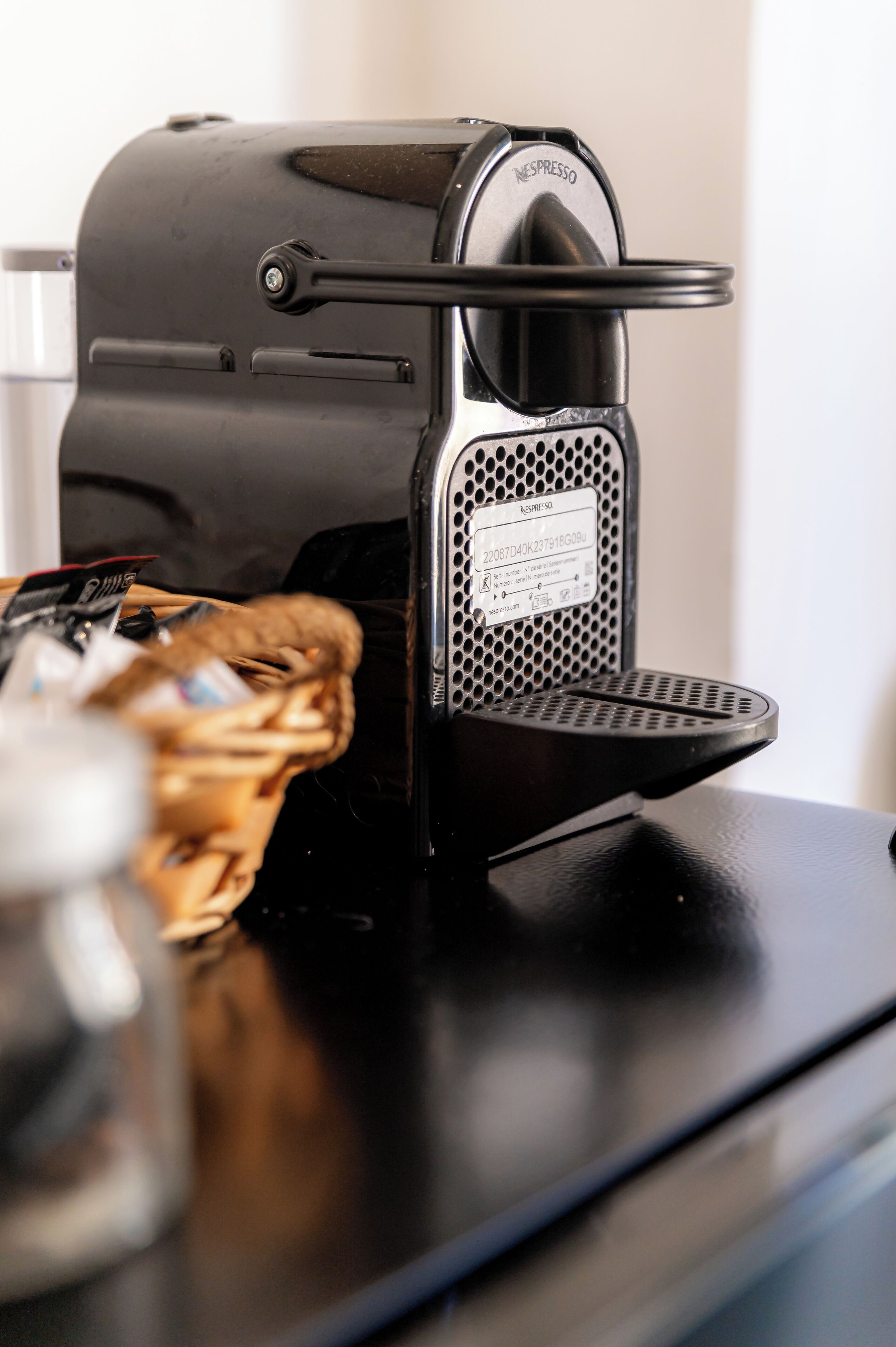 A black Nespresso coffee machine on a countertop with a basket of items and a glass.