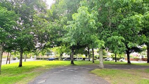 A paved pathway surrounded by lush green trees and grass, leading to a parking area with several cars.