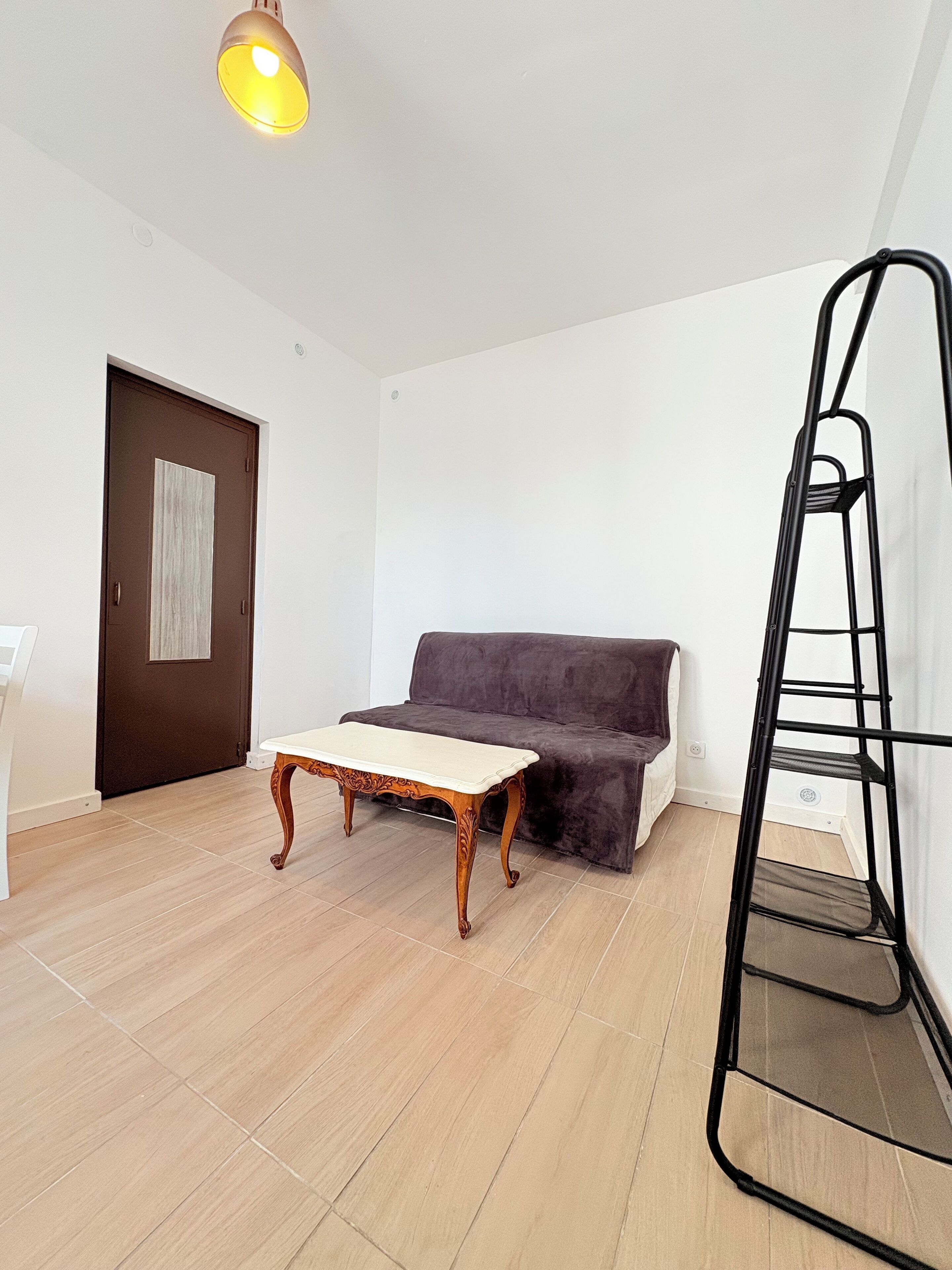 A room with a wooden floor, a brown door, a white wall, a dark brown sofa, a white table, and a black metal ladder.