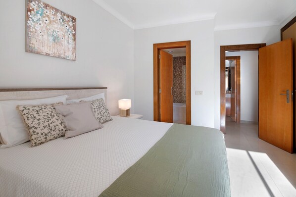 A bedroom with a bed, pillows, a nightstand, a lamp, a painting on the wall, and an open door leading to a bathroom.