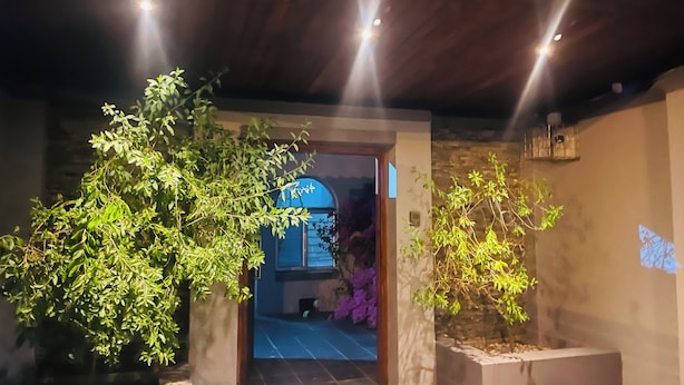 Una entrada con techo de madera, pared de piedra y puerta que da a un cuarto con flores púrpuras.