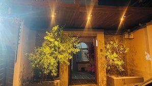 Un espacio al aire libre cubierto, con techo de madera y pared de piedra. Hay dos plantas en macetas y una puerta que da a un edificio.