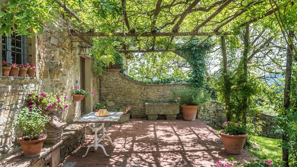A stone building with a pergola, potted plants, and a table with chairs.