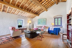 A room with a wooden ceiling, a leather sofa, a small table with books, a chair, a table with a blue tablecloth, and a bookshelf.
