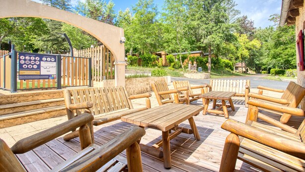 Een terras van hout buiten, met banken en tafels, omringd door bomen en een houten hek.