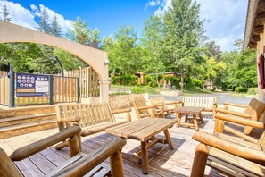 Una terraza exterior de madera con bancos y mesas, rodeada de árboles y una valla de madera.