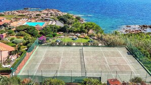 A tennis court surrounded by lush greenery and a pool, with a view of the sea and rocky coastline.