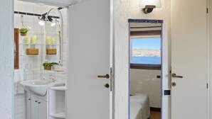 Une salle de bain avec un lavabo blanc, un miroir et des étagères contenant des articles de toilette. Une porte donne sur une chambre à coucher avec un lit et une vue sur la mer.