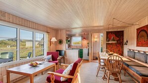 Acogedor interior de cabaña con paredes y techo de madera, comedor con mesa y sillas, cocina con pileta y alacenas, y ventanas amplias que ofrecen vistas al exterior.