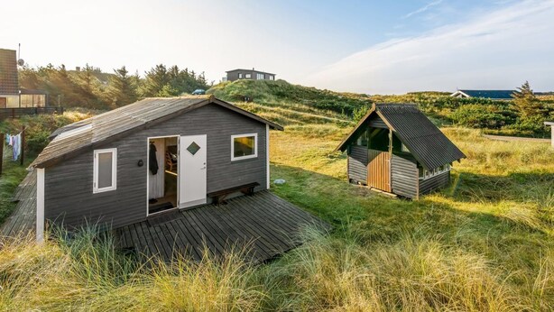 Una cabaña moderna con una terraza de madera, ubicada en una zona cubierta de hierba con vista al mar y otros edificios al fondo.