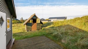 Una terraza de madera que lleva a una cabaña pequeña con techo inclinado, rodeada de campos verdes y un cielo despejado.