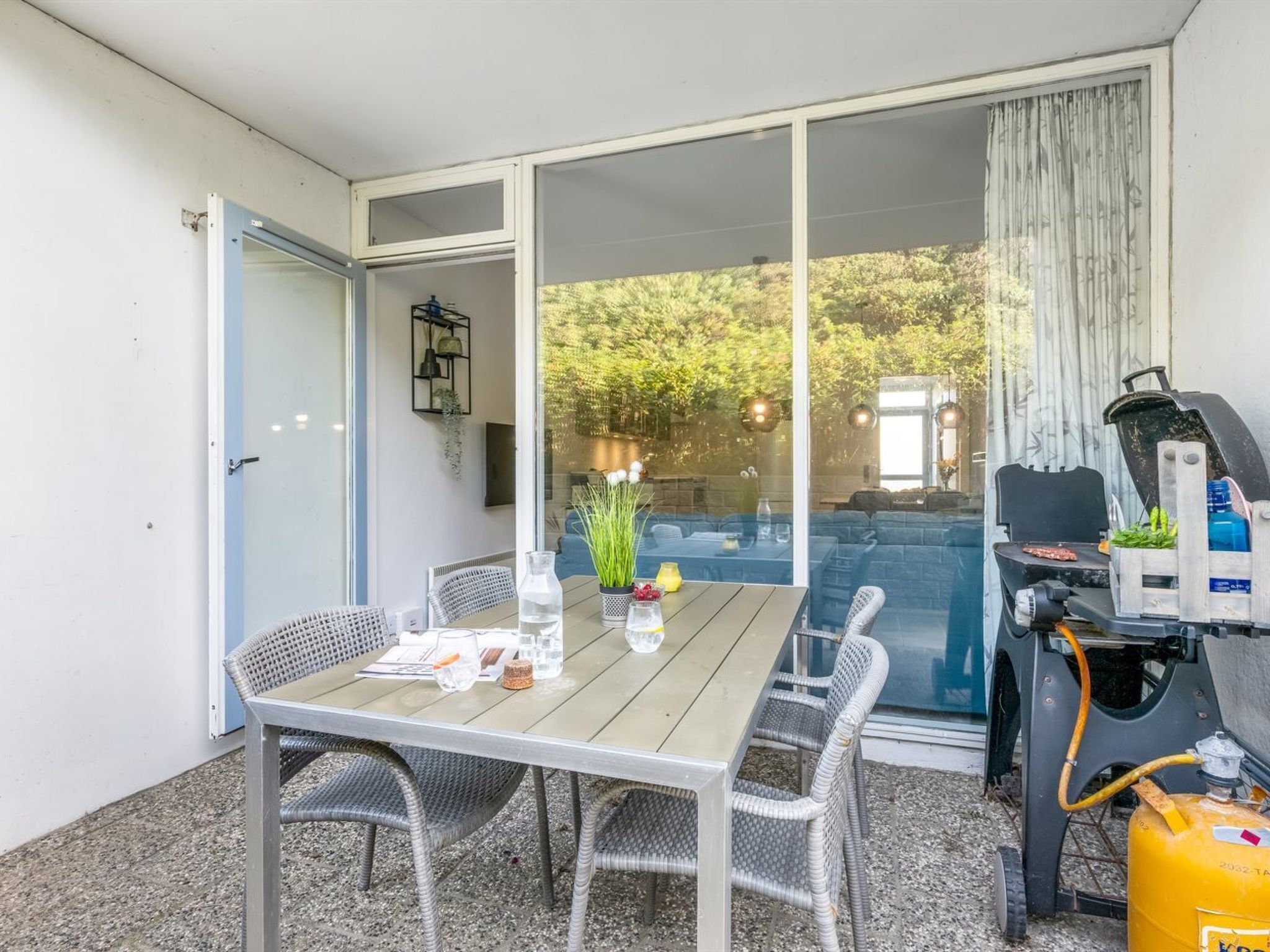 An outdoor dining area with a table and chairs, a grill, and a sliding glass door leading to a garden.