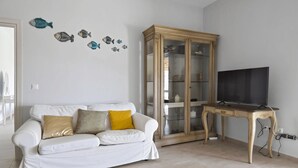 A living room with a white sofa, a wooden cabinet with glass doors, and a television on a wooden table. - Clio Apartment Kea (Otzias)