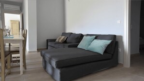 A modern living room with a dark grey sofa, two pillows, a dining table with chairs, and a kitchen area in the background. - Clio Apartment Kea (Otzias)