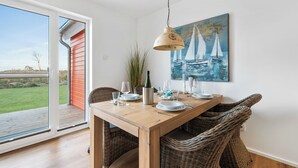Uma sala de jantar com uma mesa de madeira posta para quatro pessoas, um quadro de barcos à vela na parede e vista para um deck externo através de uma porta de vidro deslizante.