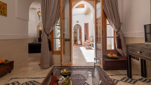 A room with a view of a courtyard, a dining table with a tray of food, a bottle, and a television on a stand. - Entire Medina Riad with Rooftop & pool (Marrakesh)