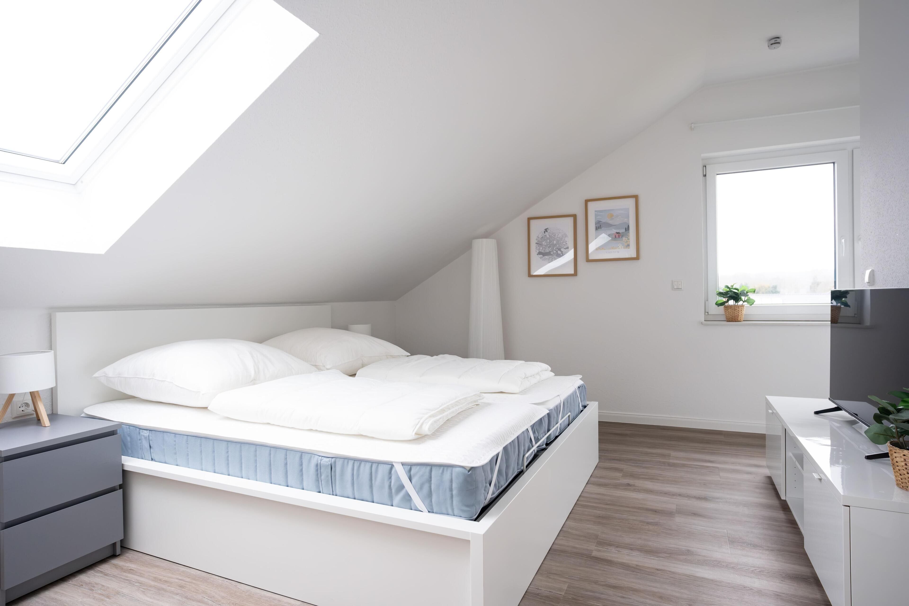A modern bedroom with a skylight, a bed with white bedding, a bedside table, a desk, and a window with a plant.