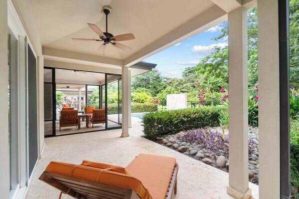 Un espace extérieur abrité, doté d’un ventilateur de plafond, d’un coin salon avec des coussins orange et d’une vue sur un jardin avec une piscine.