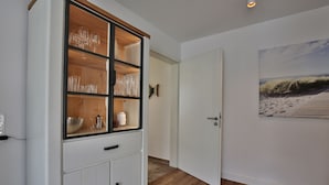 A hallway with a white cabinet containing glassware and a painting of a beach scene on the wall. - Seeblick Nordlicht - Comfortable accommodation on Wangerooge with terrace and garden (Wangerooge)