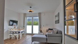 A modern living room with a sofa, a TV, a dining table with chairs, and a large window with curtains. - Seeblick Nordlicht - Comfortable accommodation on Wangerooge with terrace and garden (Wangerooge)