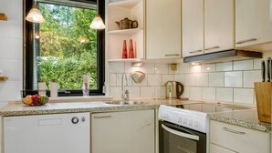 A compact kitchen with white cabinets, a dishwasher, an oven, and a window with a view of greenery.