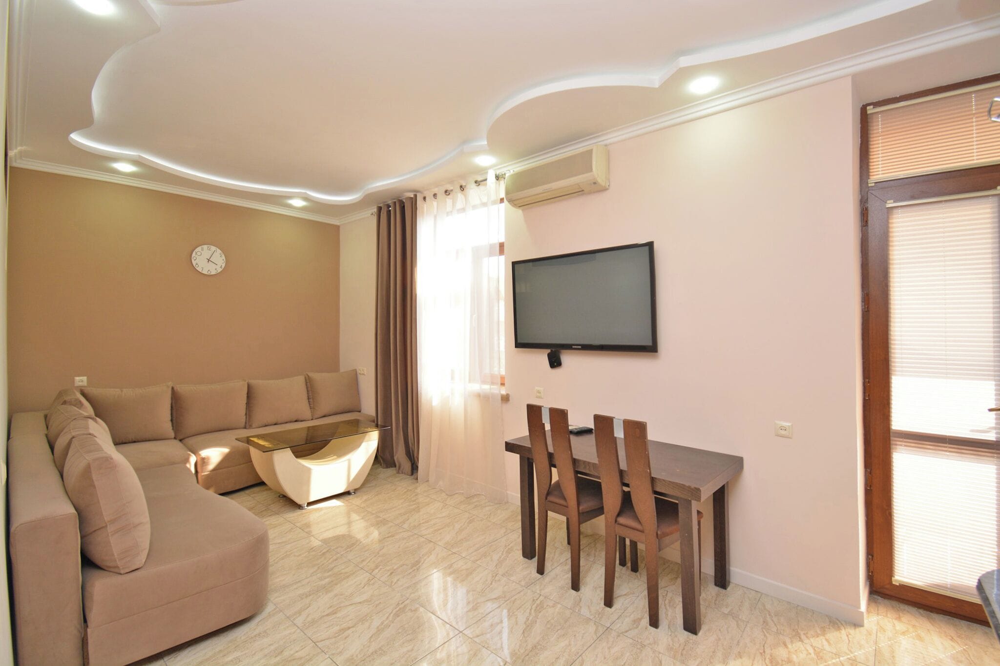 A living room with a beige sofa, a dining table with chairs, a flat-screen TV mounted on the wall, and a window with curtains.