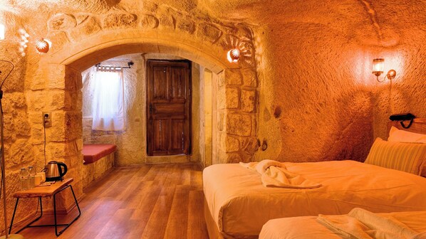 A rustic stone-walled room with a wooden door, a bench, a bed, and a small table with a kettle and bottles. - Anjelic Cave Cappadocia (Ürgüp)