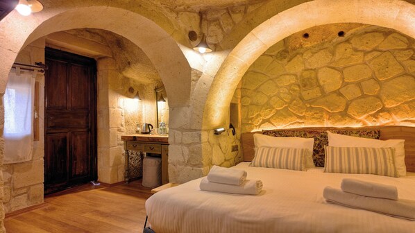 Une chambre avec des murs en pierre, une porte en bois, une table de chevet avec une bouilloire, et un lit avec des oreillers et des serviettes.